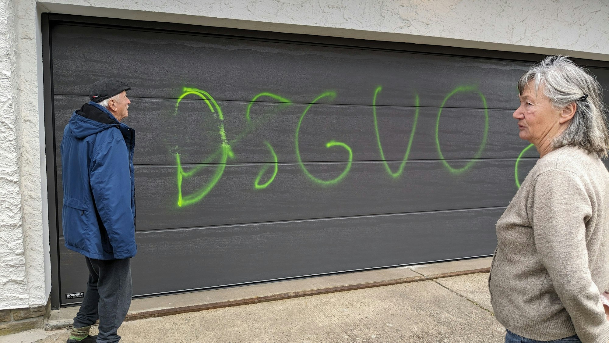 Peter und Rosemarie Leister stehen vor ihrem mit grüner Farbe verunzierten Garagentor. Die Aufschrift lautet DSGVO.