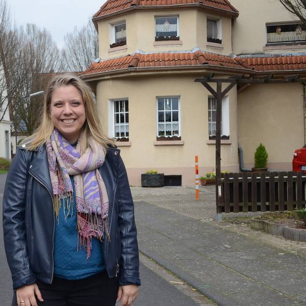 Eine junge Frau mit blonden Haaren auf einer Straße. Hinter ihr sind ältere Häuser einer Siedlung zu sehen.