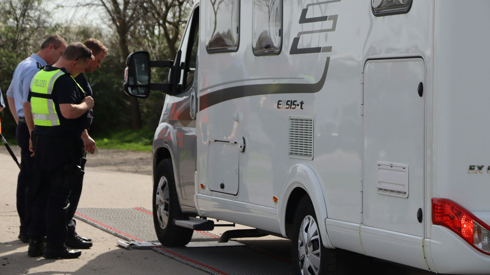 Ein Wohnmobil steht mit dem linken Vorderrad auf einer Waage. Drei Polizeibeamte lesen das Gewicht ab.