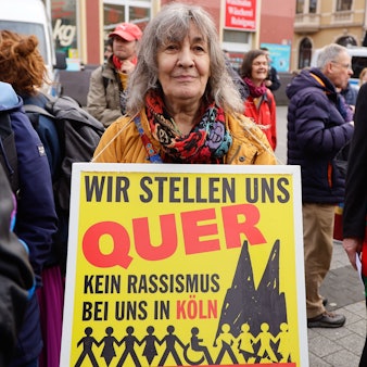 Menschen bei einer Demonstration. Eine Frau trägt ein Plakat mit der Aufschrift: „Wir stellen quer - Kein Rassismus bei uns in Köln“.