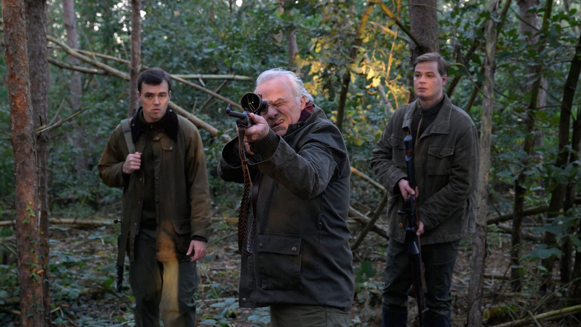 Die drei Charaktere stehen im Wald. Dr. Albrecht Richtmann, der Chef der Kanzlei, zielt mit einem Jagdgewehr. Er steht mittig zwischen seinem Sohn Konstantin Richtmann und Daniel Pillokat, die beide der gleichen Anwaltskanzlei des Schützen arbeiten.