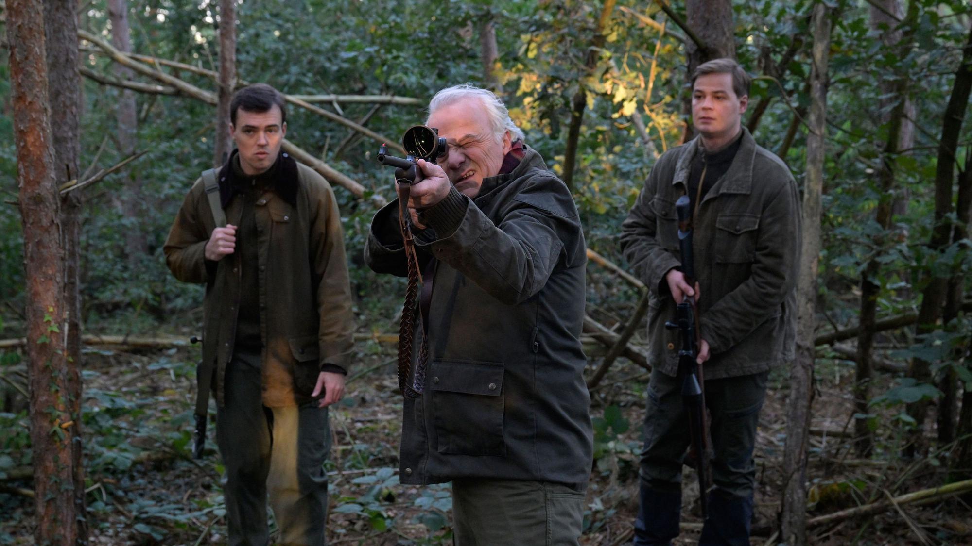 Die drei Charaktere stehen im Wald. Dr. Albrecht Richtmann, der Chef der Kanzlei, zielt mit einem Jagdgewehr. Er steht mittig zwischen seinem Sohn Konstantin Richtmann und Daniel Pillokat, die beide der gleichen Anwaltskanzlei des Schützen arbeiten.