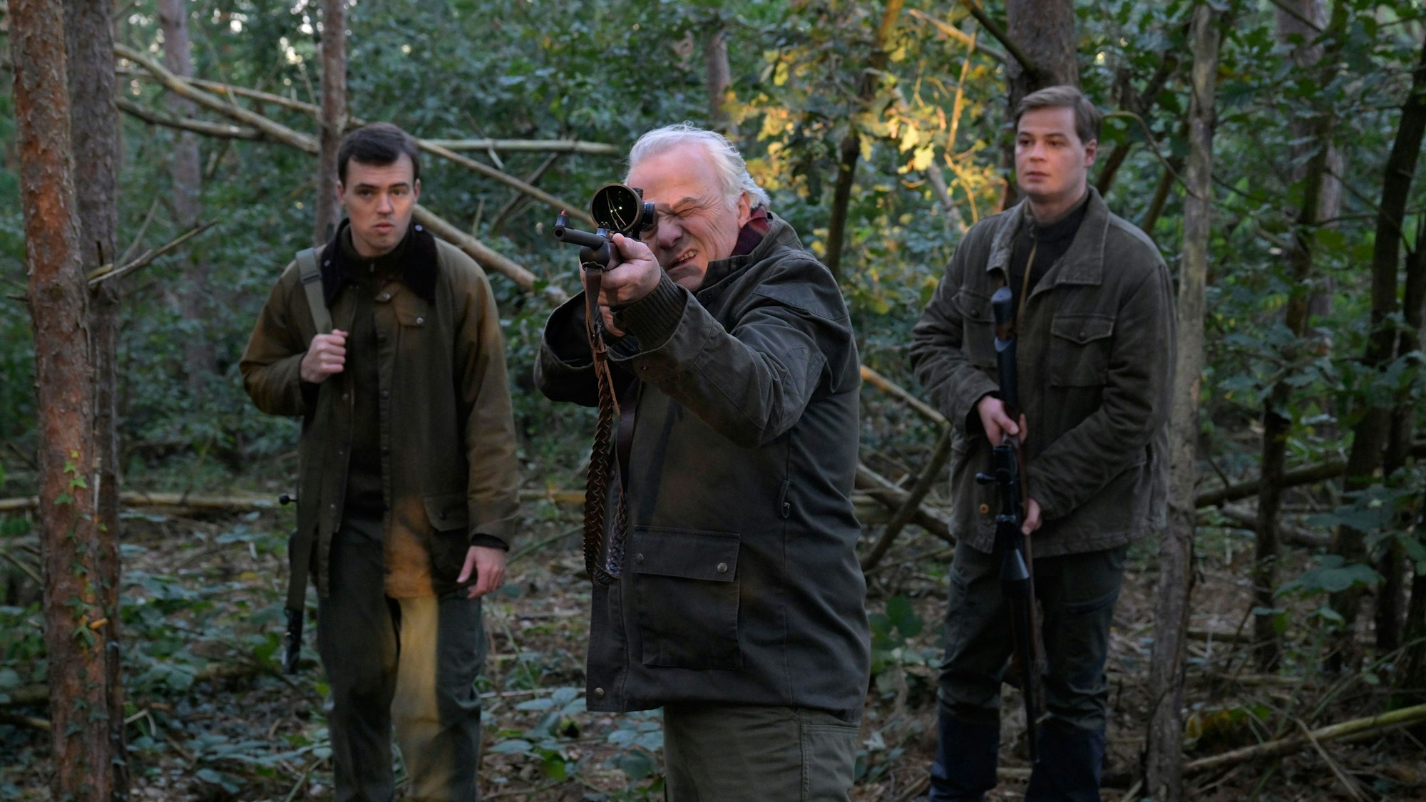 Die drei Charaktere stehen im Wald. Dr. Albrecht Richtmann, der Chef der Kanzlei, zielt mit einem Jagdgewehr. Er steht mittig zwischen seinem Sohn Konstantin Richtmann und Daniel Pillokat, die beide der gleichen Anwaltskanzlei des Schützen arbeiten.