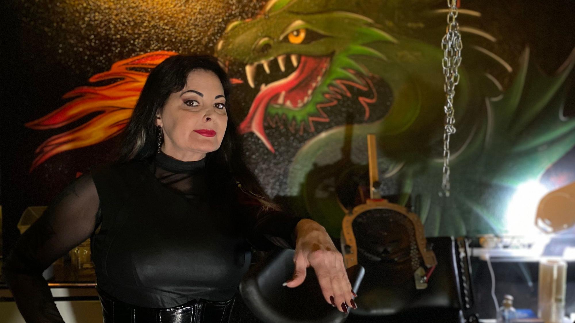 Die Domina steht in ihrem Studio. Ein Drache ist auf die Wand im Hintergrund gemalt.