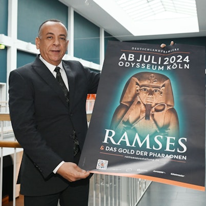 Mostafa Waziri hält in den Gängen des Odysseums Köln ein Plakat der Ausstellung "Ramses & Das Gold der Pharaonen". Die Ausstellung ist ab 13. Juli 2024 zu Gast im Odysseum. Dr. Mostafa Waziri hat sich vor Ort ein Bild gemacht.