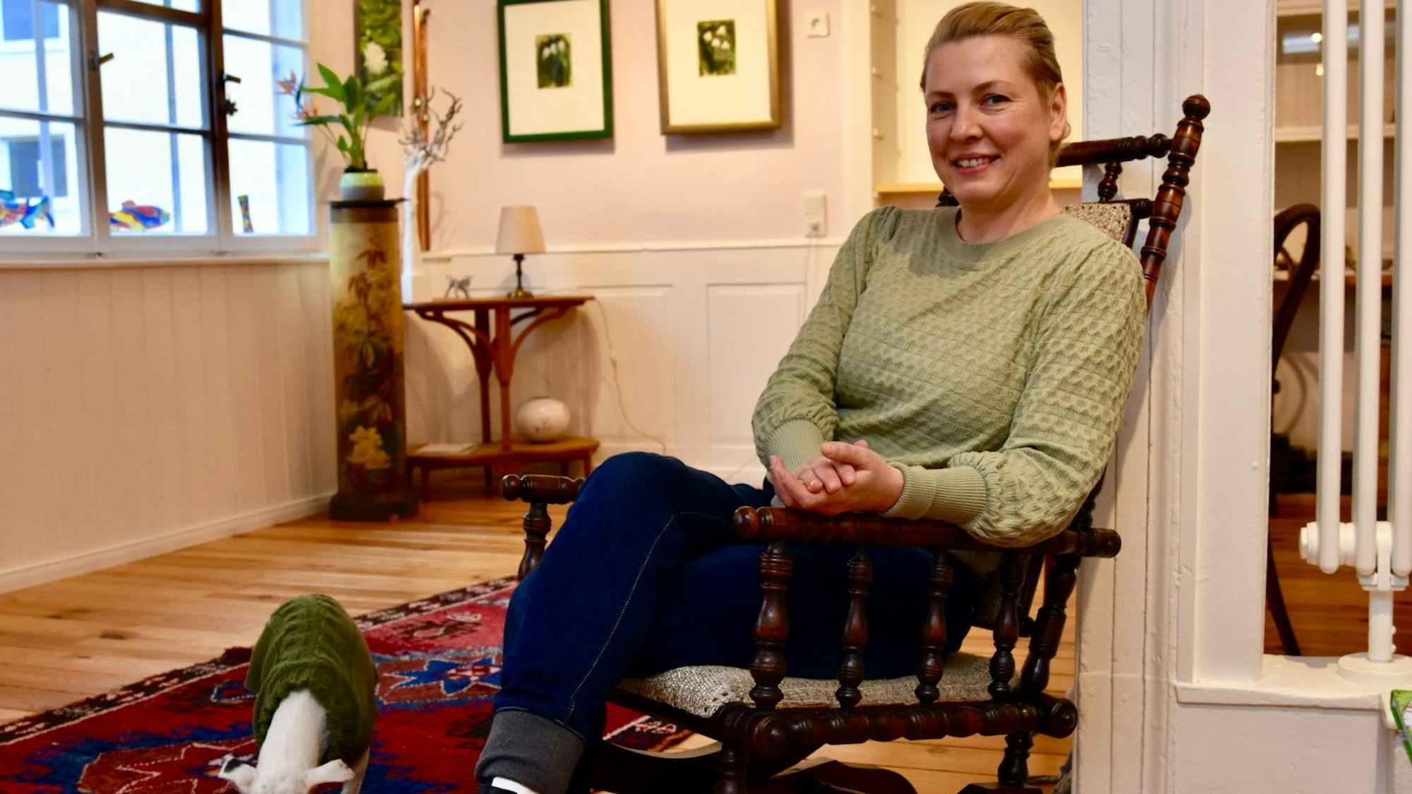 Eine Frau in einem Schaukelstuhl. Auf dem Boden steht ein kleiner weißer Hund mit einem grünen Wollpullover.