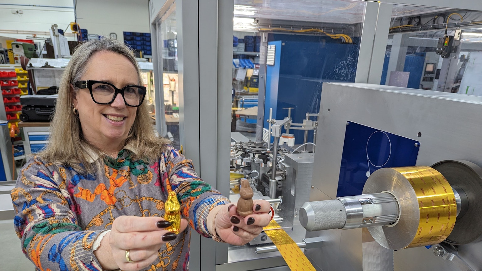 Eine Frau steht vor einer großen Maschine und zeigt jeweils einen Schokohasen mit und ohne Einwicklung.