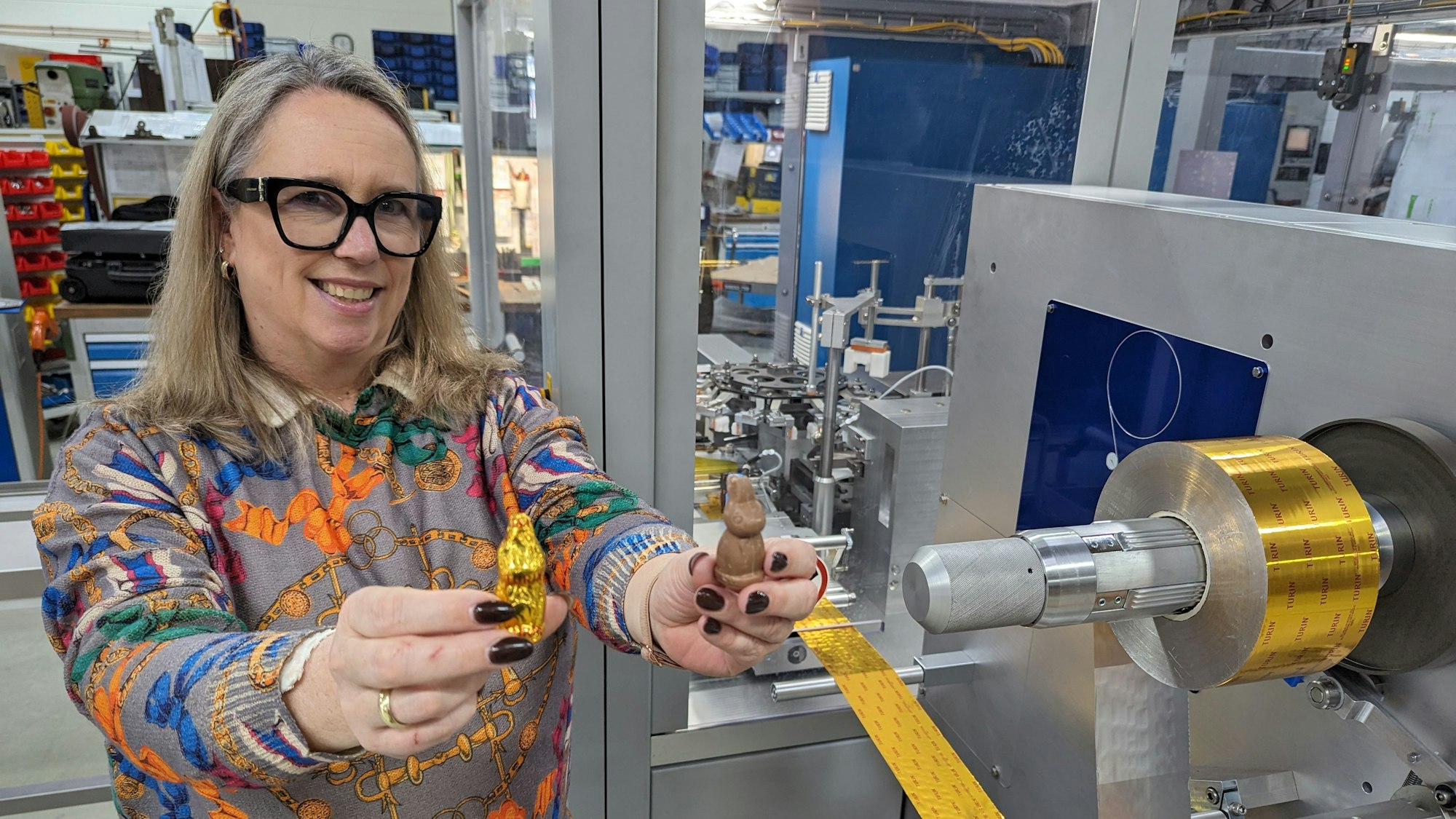 Eine Frau steht vor einer großen Maschine und zeigt jeweils einen Schokohasen mit und ohne Einwicklung.