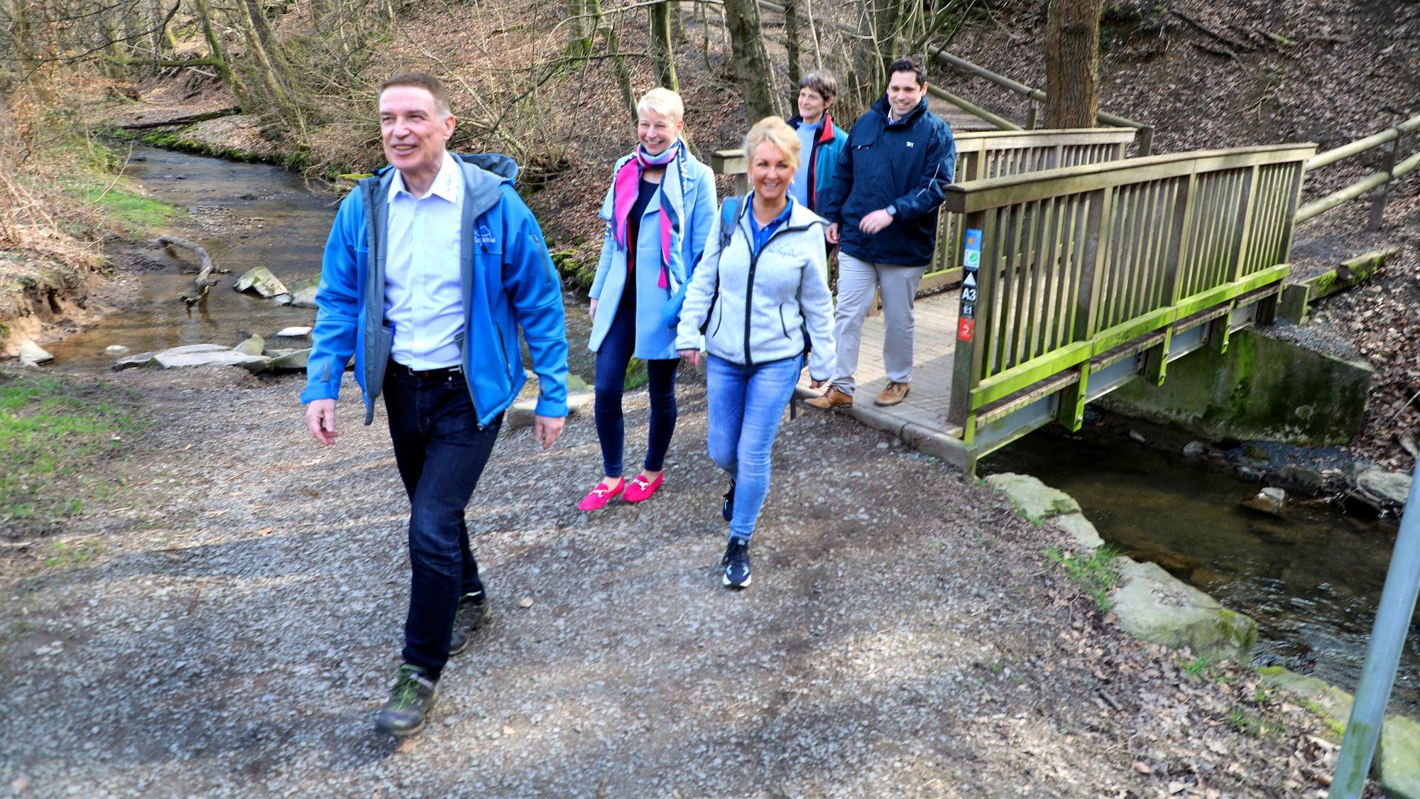 Die verantwortlichen Projektträger des Bergischen Wanderbusses von Kreis, Kommunen und Regionalverkehr Köln GmbH wandern auf einem Weg, im Hintergrund sind eine Brücke und der Eifgenbach zu sehen.