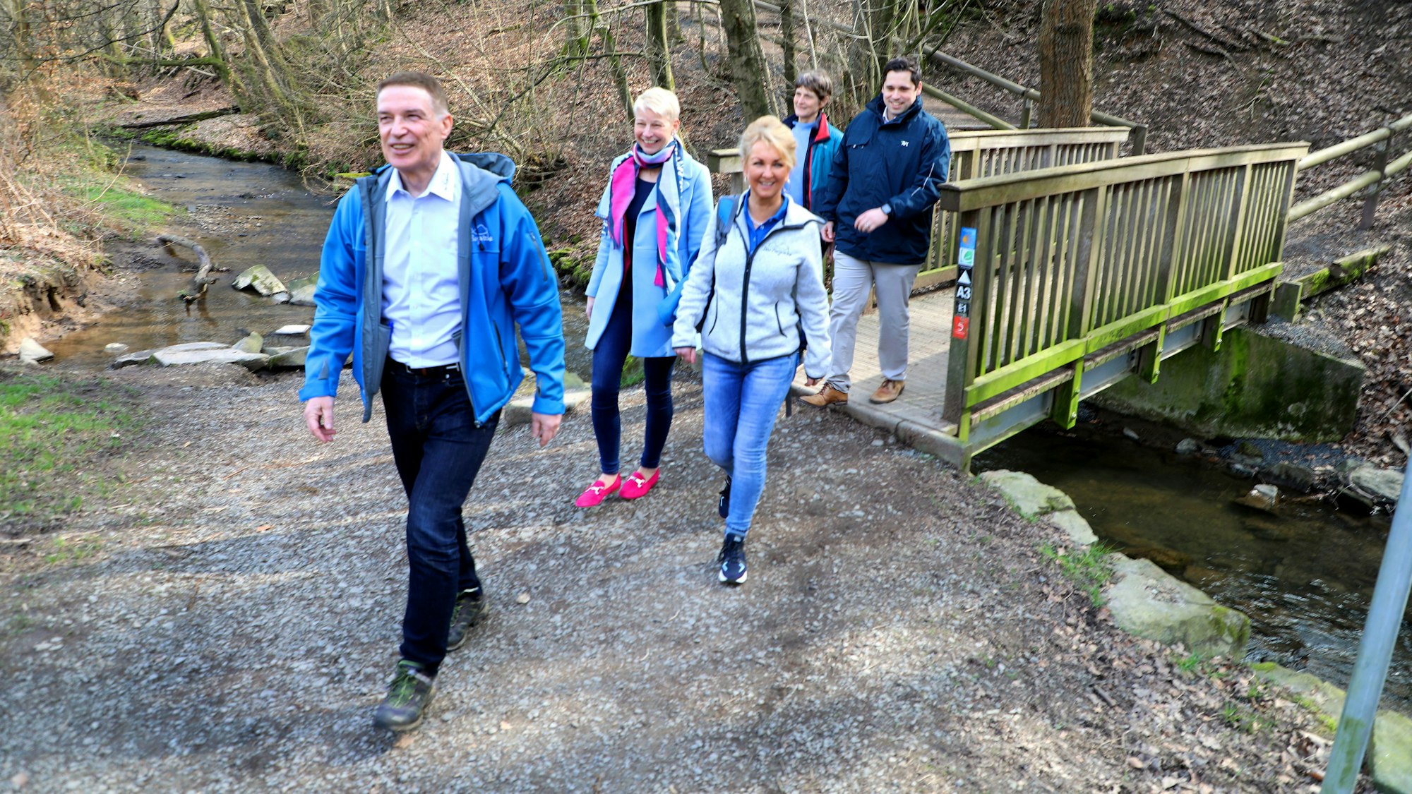 Die verantwortlichen Projektträger des Bergischen Wanderbusses von Kreis, Kommunen und Regionalverkehr Köln GmbH wandern auf einem Weg, im Hintergrund sind eine Brücke und der Eifgenbach zu sehen.