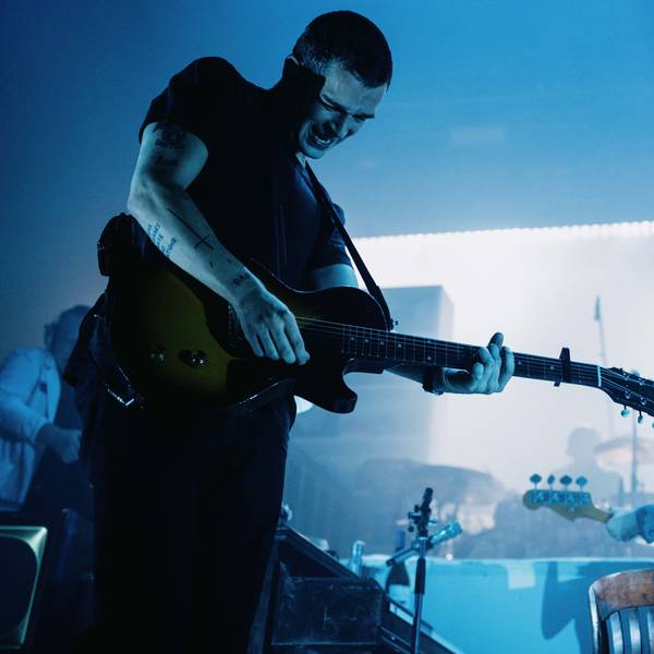 Matty Healy hat kurze Haare. Er spielt seine elektrische Gitarre und trägt einen Ausdruck von starker Anstrengung. Die Bühne ist bläulich erleuchtet.