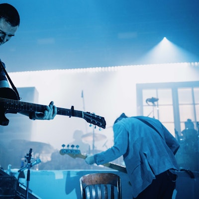 Matty Healy hat kurze Haare. Er spielt seine elektrische Gitarre und trägt einen Ausdruck von starker Anstrengung. Die Bühne ist bläulich erleuchtet.