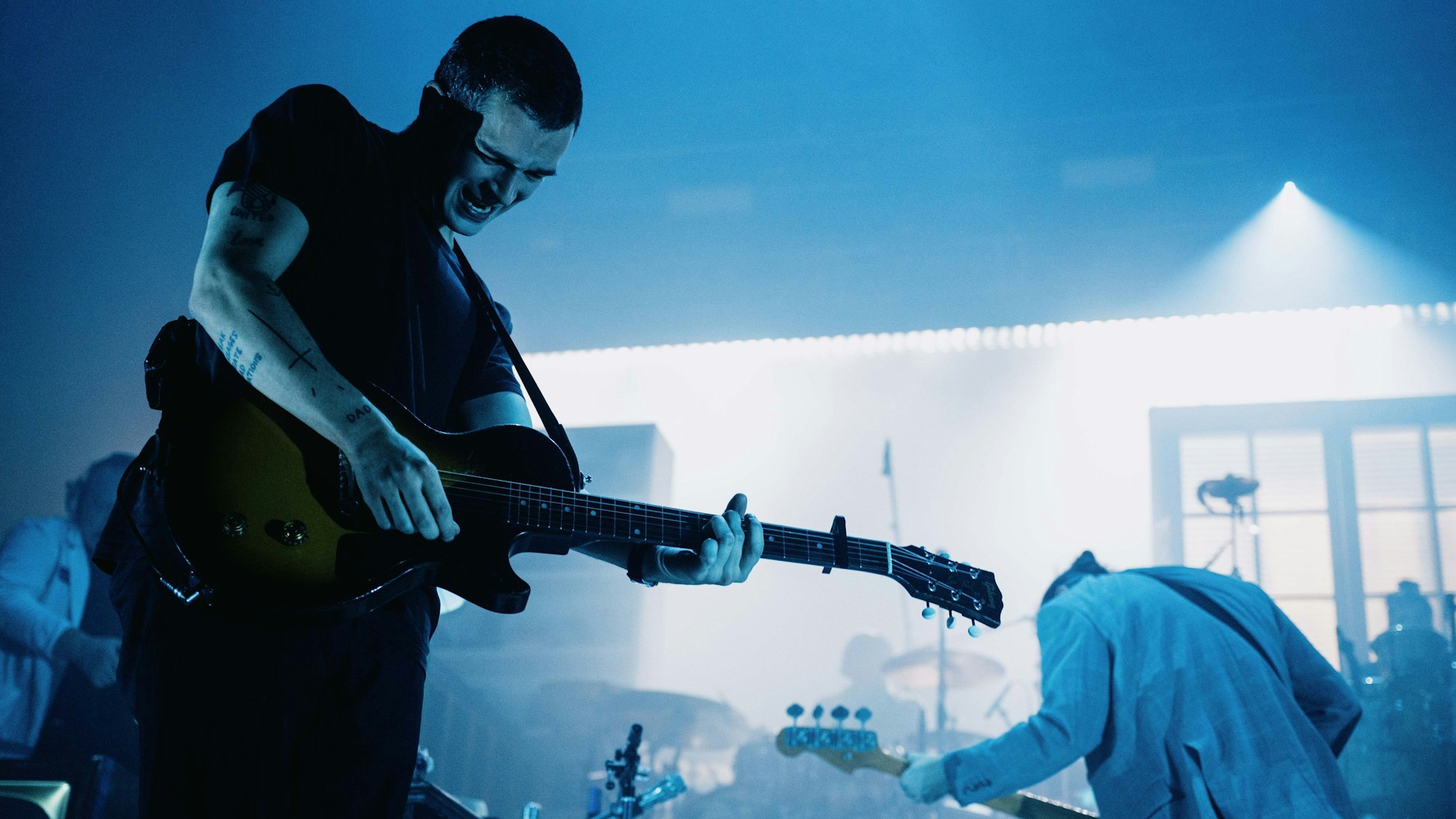 Matty Healy hat kurze Haare. Er spielt seine elektrische Gitarre und trägt einen Ausdruck von starker Anstrengung. Die Bühne ist bläulich erleuchtet.