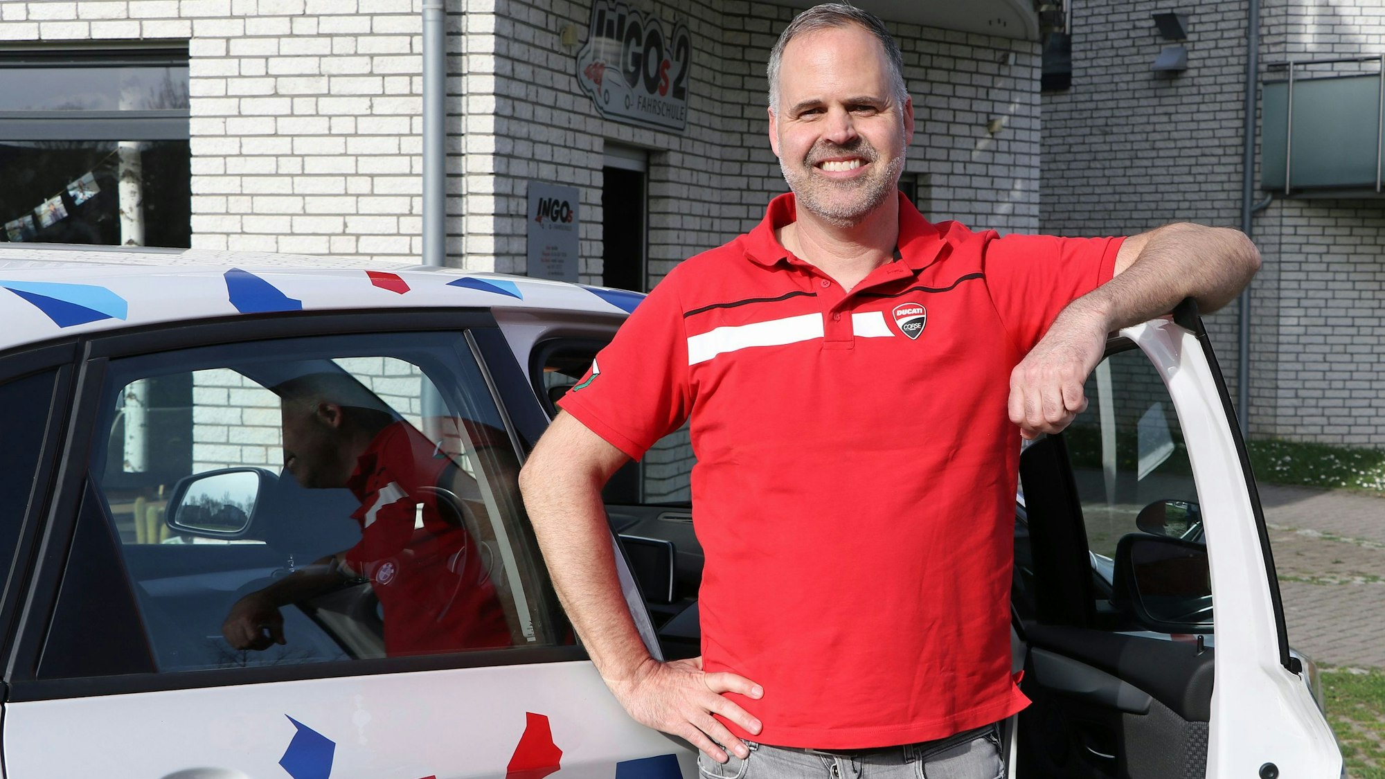 Ein Mann in rotem T-Shirt steht an einer geöffneten Autotür und lächelt in die Kamera.