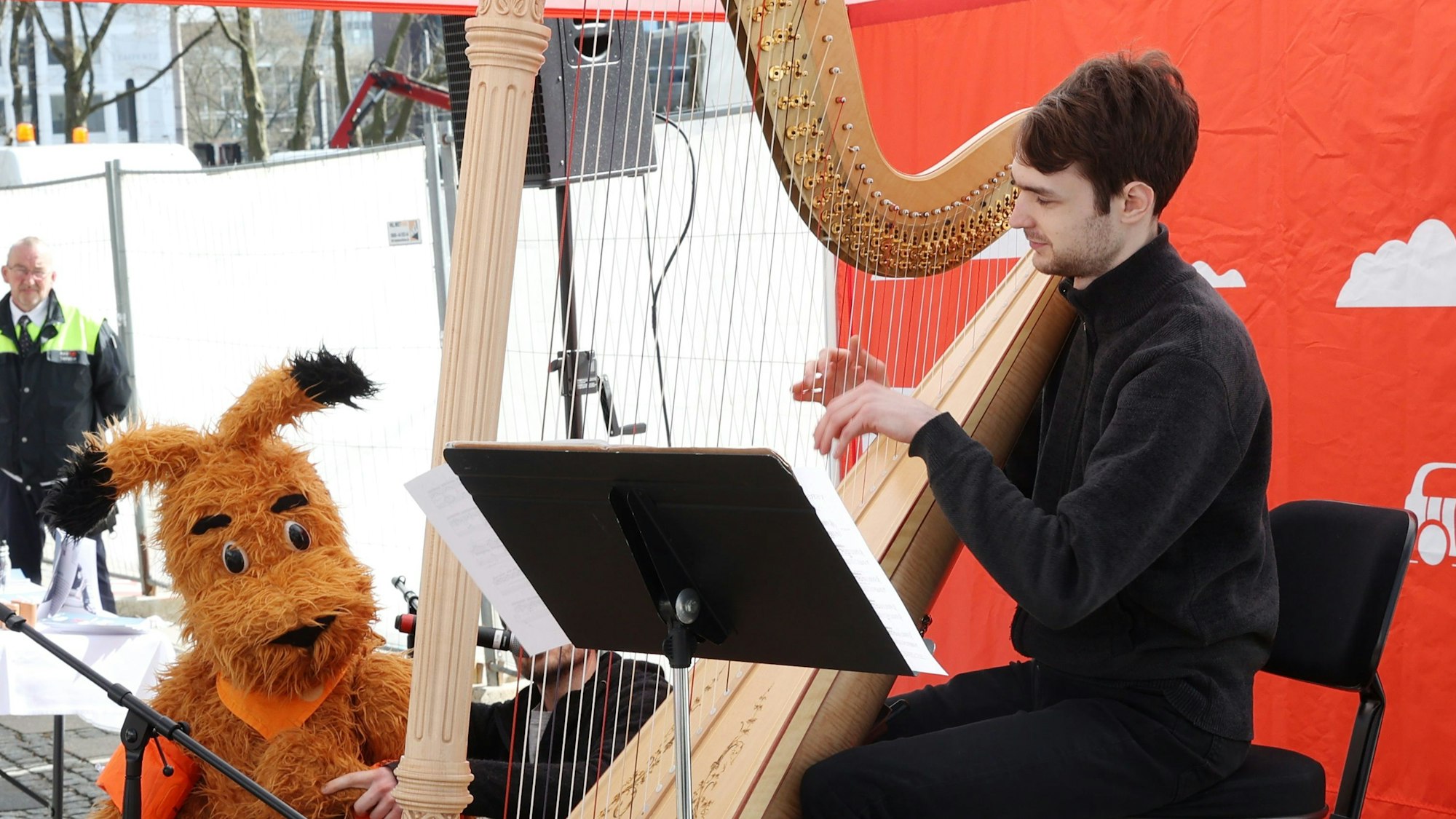 Zu sehen ist Mann, der auf einer Harfe spielt und das Maskottchen des Gürzenich-Orchesters.