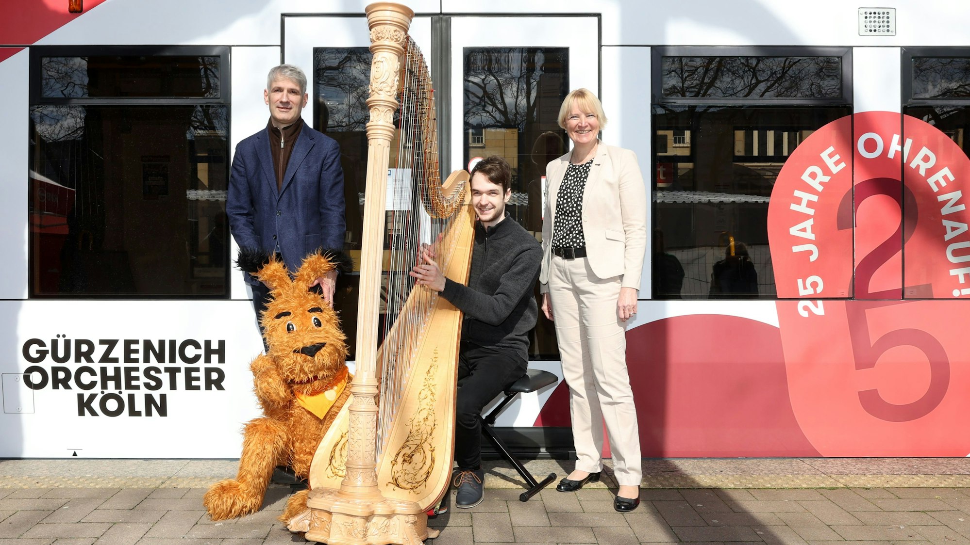 Zu sehen sind drei Menschen, die sich neben einer KVB-Stadtbahn befinden. Einer sitzt an einer Harfe.