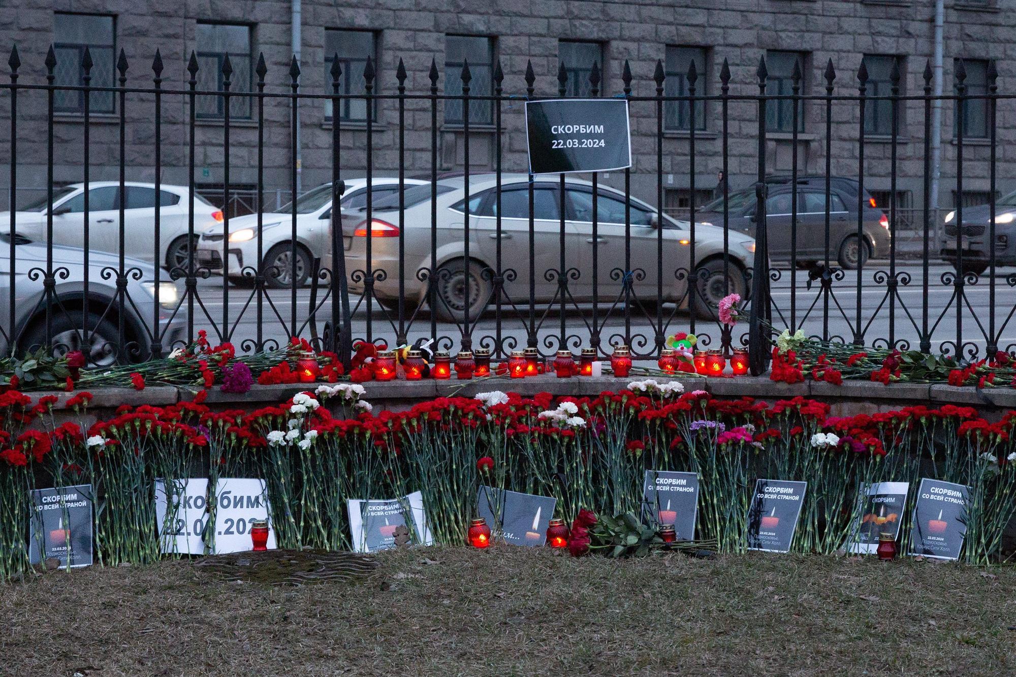 Vor einem Zaun, der ein Grundstück von einer befahrenen Straße trennt, liegen Blumen und Gedenkkerzen mit Bildern.