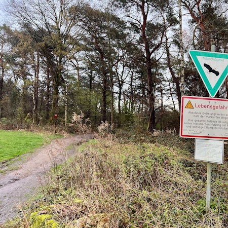 Ein Schild mit dem Hinweis „Lebensgefahr“ in der Wahner Heide weist darauf hin, dass Flächen außerhalb der markierten Wege nicht betreten werden dürfen. Im Hintergrund ist ein Wald zu sehen.
