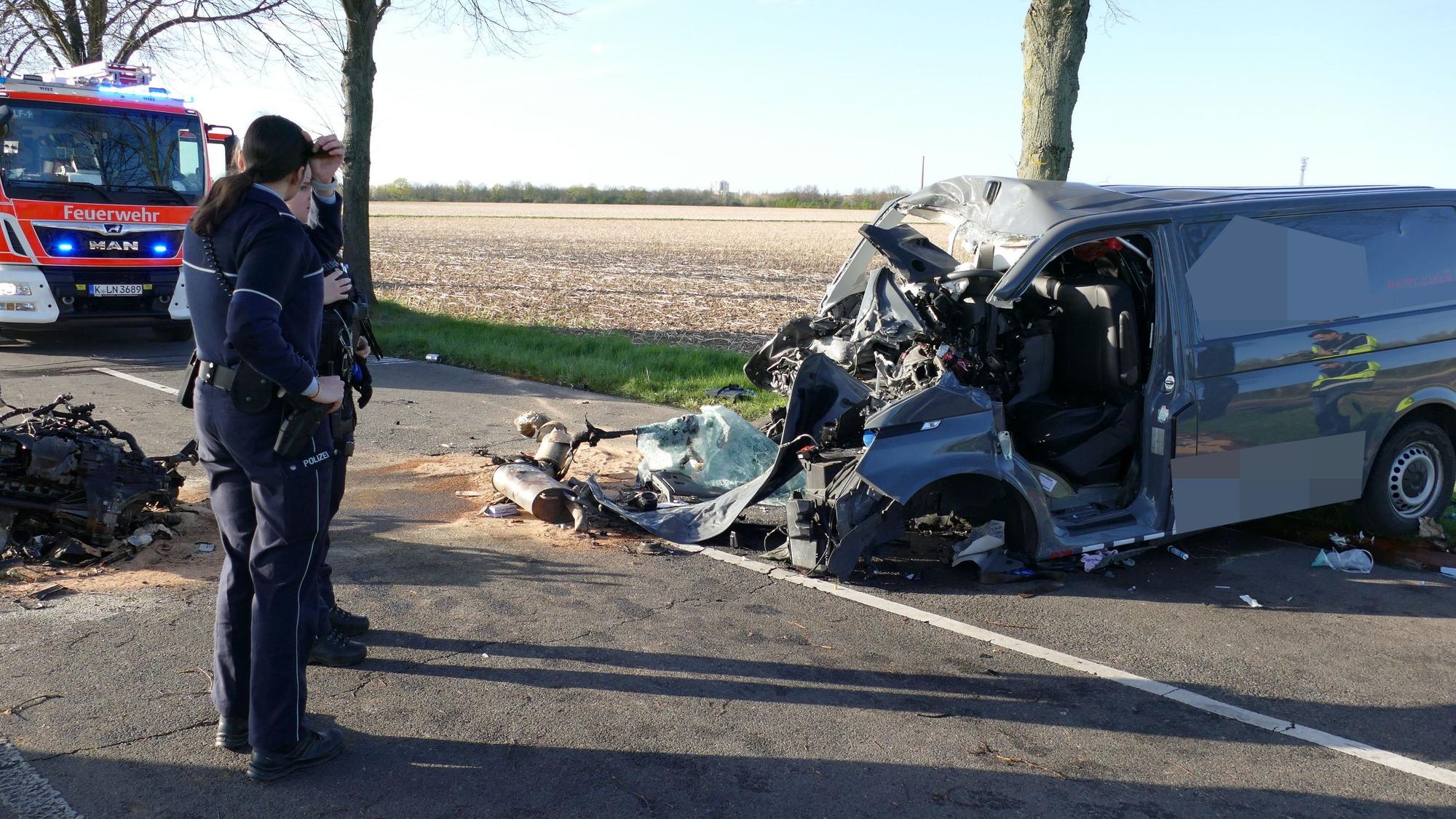 Zu sehen ist ein zerstörtes Fahrzeug auf einer Landstraße, sowie ein Feuerwehrwagen und zwei Personen.