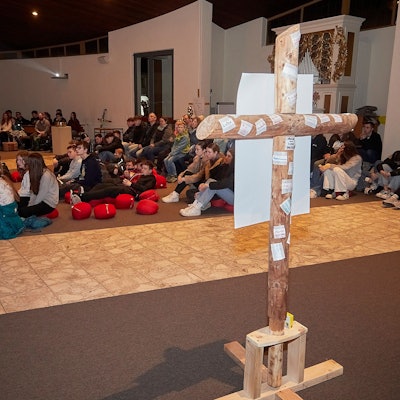 Die Teilnehmer sitzen auf Sitzkissen und auf Stühlen. Im Vordergrund steht ein Kreuz.