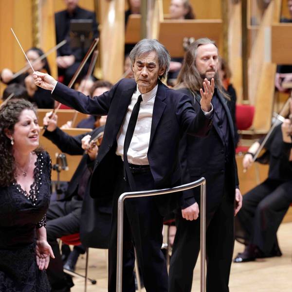 Der dirigent steht auf der Bühne der Philharmonie und breitet die Arme leicht aus, er hält einen Taktstock in de rrechten Hand. An seiner Seite sind die Solisten, im Hintergrund das Orchester.