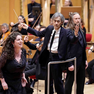 Der dirigent steht auf der Bühne der Philharmonie und breitet die Arme leicht aus, er hält einen Taktstock in de rrechten Hand. An seiner Seite sind die Solisten, im Hintergrund das Orchester.
