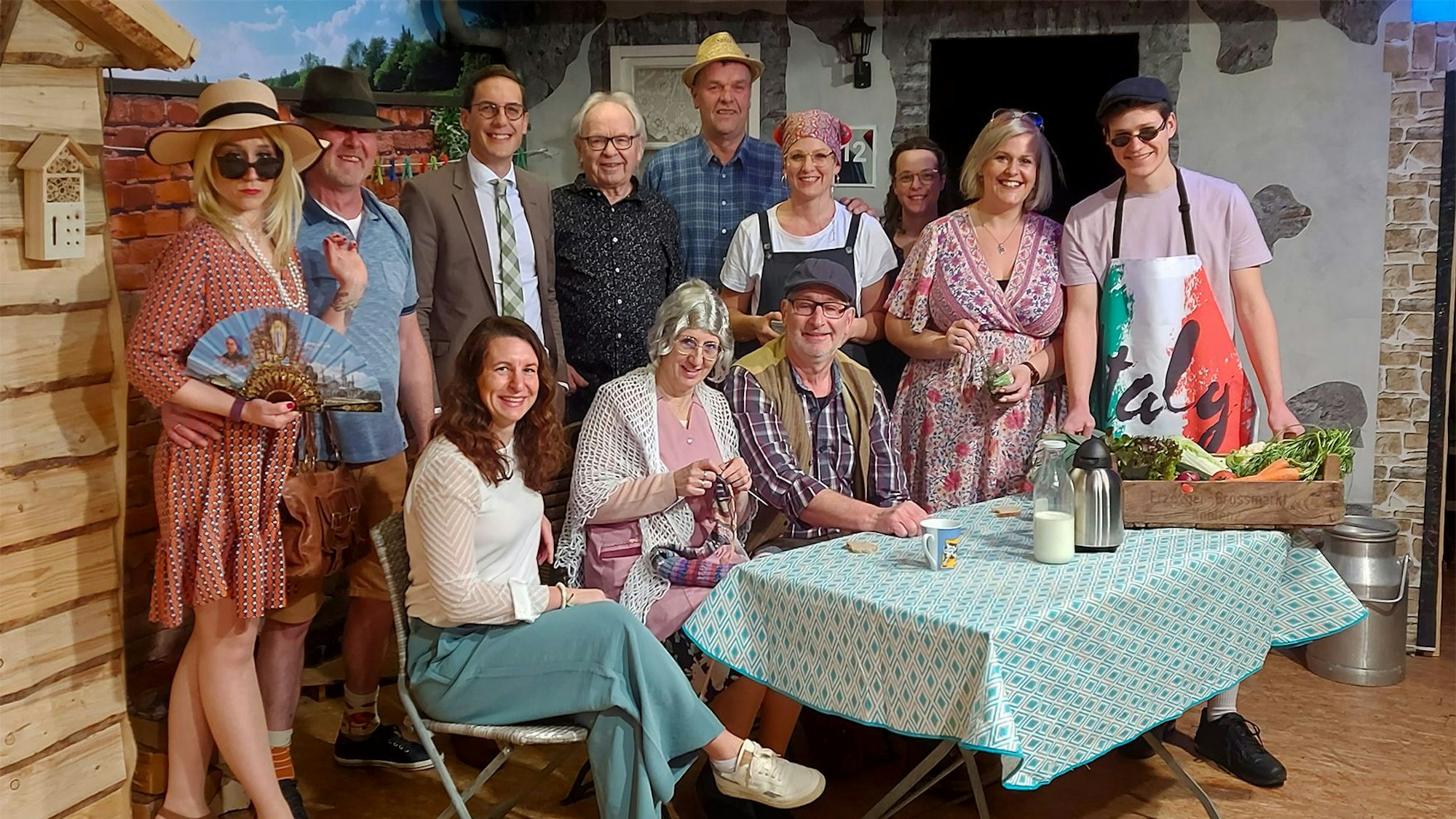 Die Schauspieler sitzen und stehen hinter einem Tisch mit Requisiten.