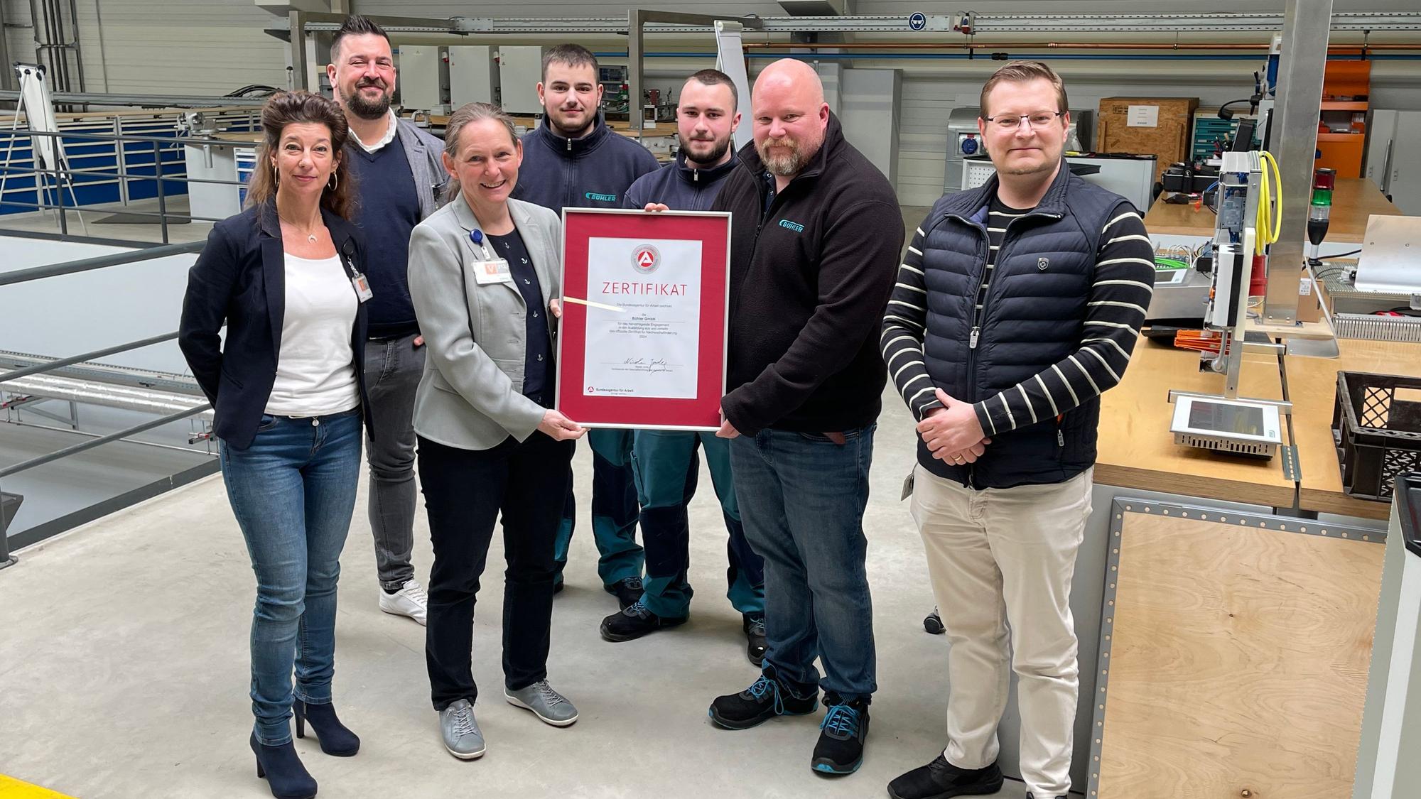 Eine Gruppe Männer und Frauen steht mit einem Bilderrahmen und dem Ausbildungszertifikat in einer Produktionshalle eines Unternehmens.