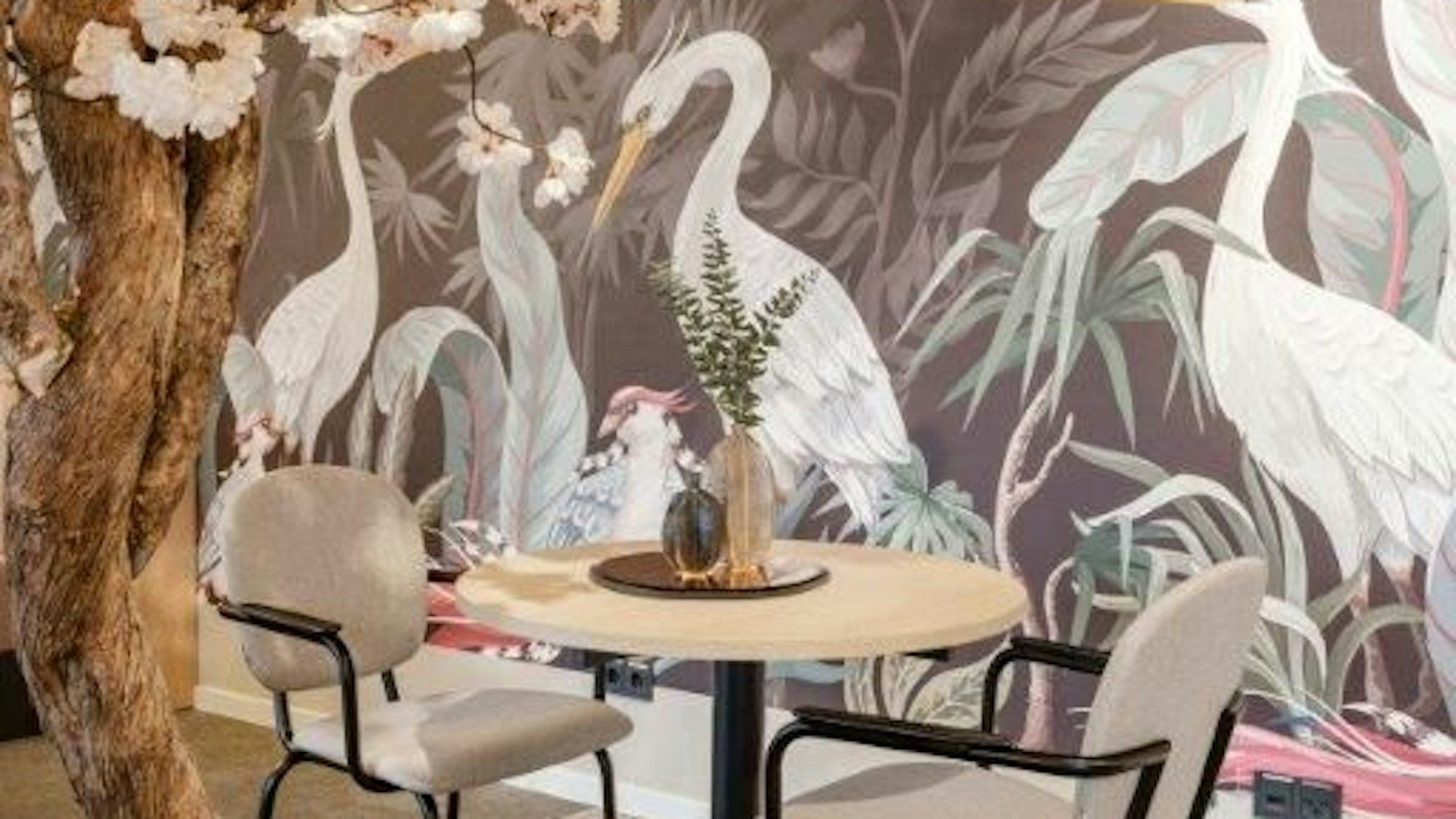 Zu sehen ist ein Tisch in einem Café mit Vogeltapete und einem Baum mit rosafarbenen Blüten.