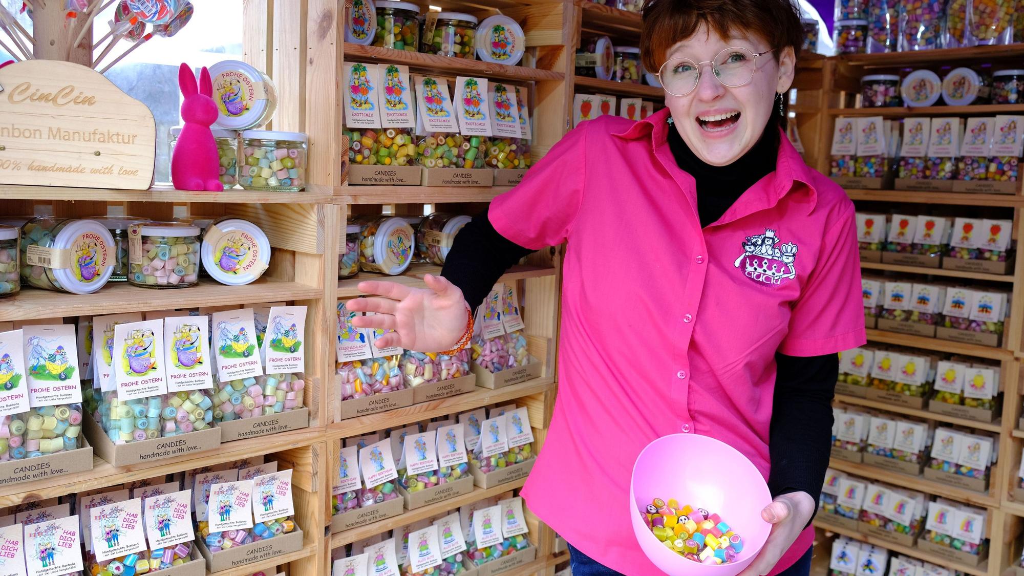 Eine junge Frau in einem pinken Shirt steht vor Regalen, in denen sich Tüten mit Bonbons befinden. In der Hand hält sie eine Schale mit einigen Bonbons zum Probieren.