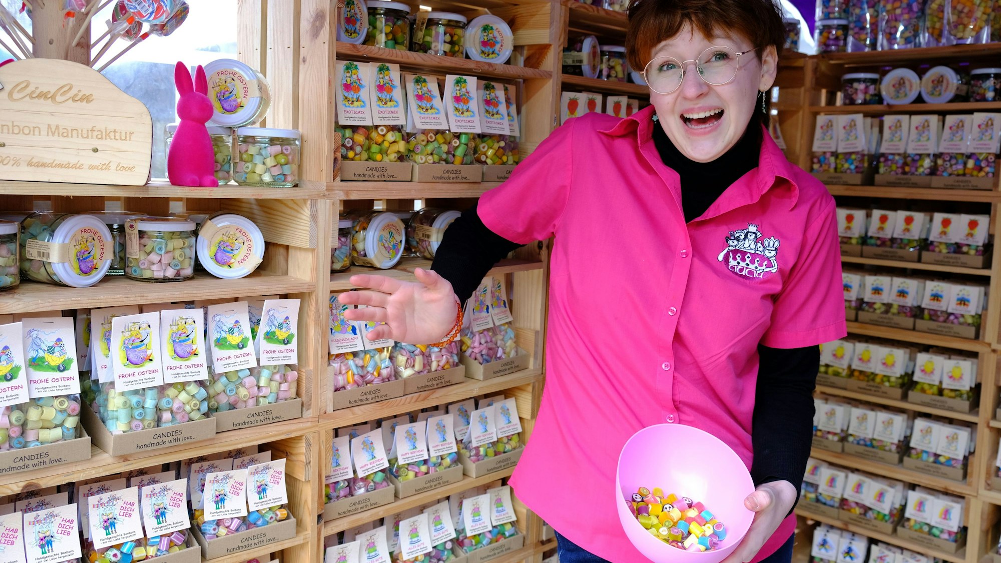 Eine junge Frau in einem pinken Shirt steht vor Regalen, in denen sich Tüten mit Bonbons befinden. In der Hand hält sie eine Schale mit einigen Bonbons zum Probieren.