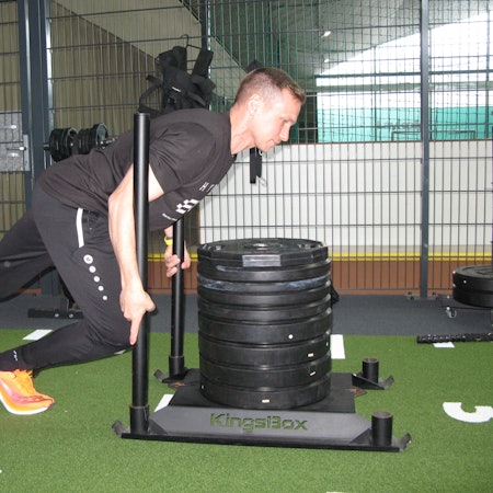 Der 38-jährige Roman Hopp, athletische Figur, schwarzes T-Shirt, schwarze Trainingshose, stemmt sich gegen einen mit großen Hantelscheiben beladenen Schlitten aus einer Metallplatte und zwei senkrecht eingelassen Stangen.