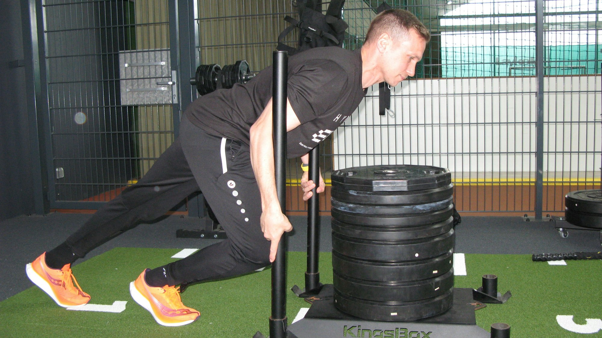 Ein Mann in Trainingskleidung schiebt eine Metallplatte, auf der sich große Hantelscheiben türmen.