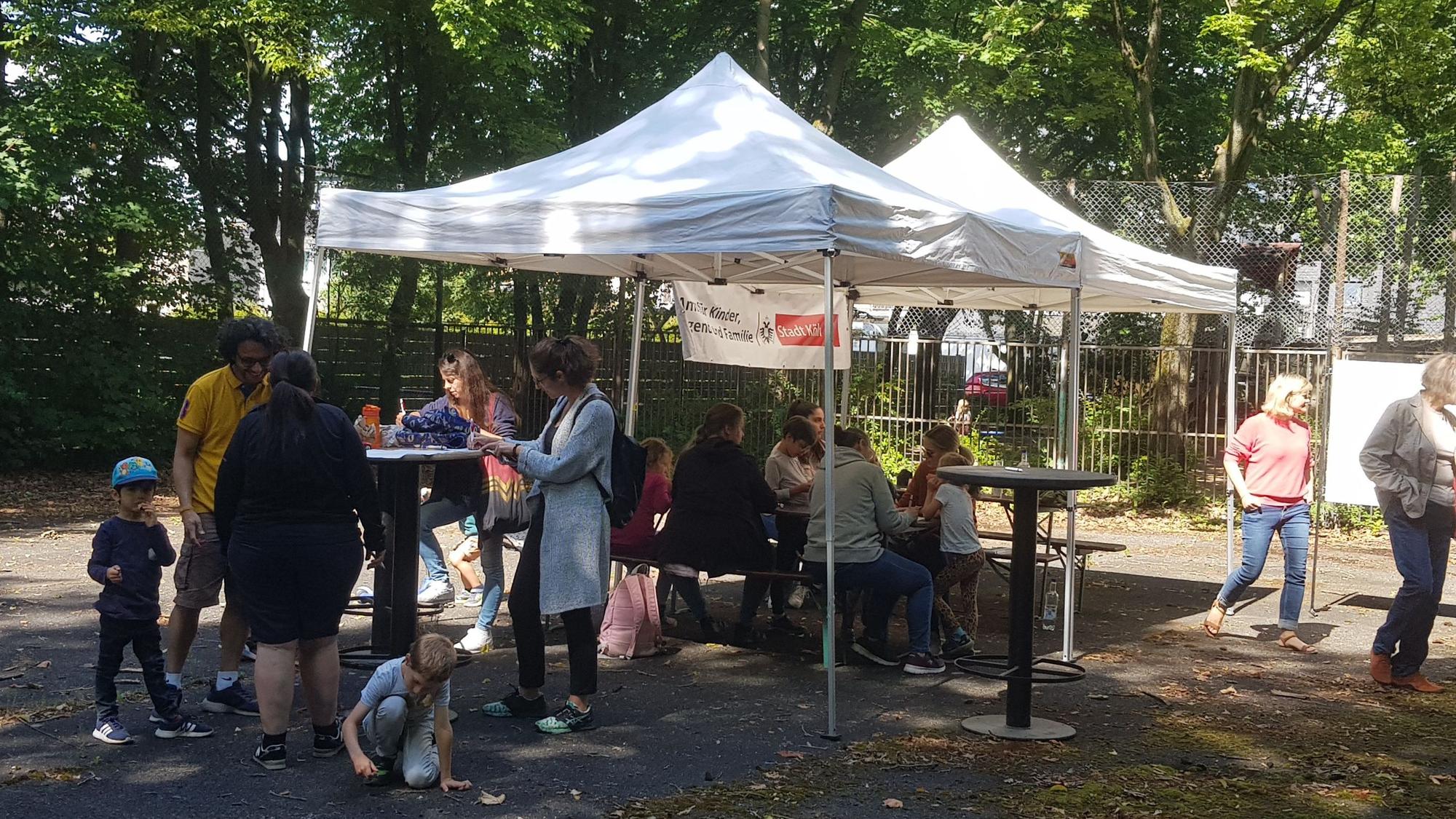 Unter einem Zelt am Bolzplatz An den Kreutzmorgen stehen und sitzen Kinder und Erwachsene.