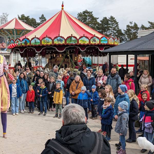 Zwei Artisten auf Stelzen unterhalten beim Jahrmarkt anno dazumal im LVR-Freilichtmuseum Kommern die Besucher. Im Hintergrund sind zwei alte Karussells zu sehen.