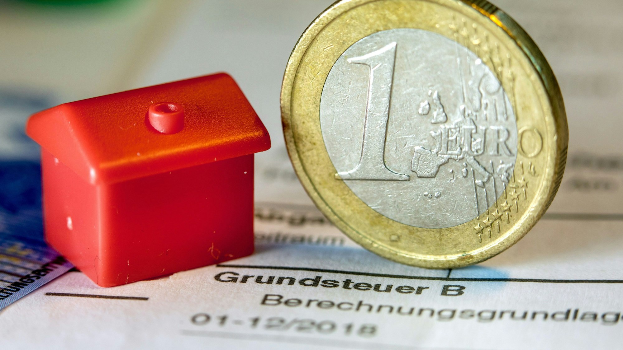 Eine Euro-Geldmünze sowie ein Spielzeughaus stehen auf einem Abgabenbescheid für die Entrichtung der Grundsteuer.