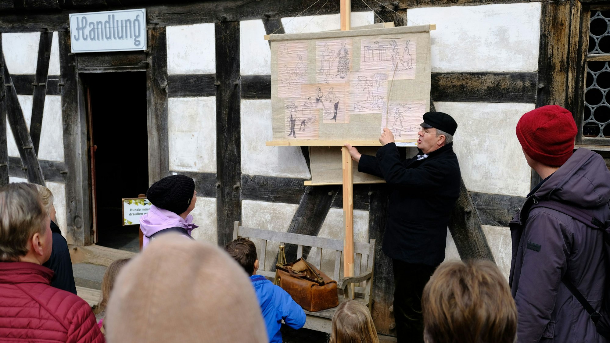 Ein Moritatensänger mit schwarzer Jacke und schwarzer Kappe steht vor einem Fachwerkgebäude im LVR-Freilichtmuseum Kommern. Er weist mit einem Stock auf eine Tafel mit verschiedenen Abbildungen, um ihn herum stehen einige Museumsbesucher.