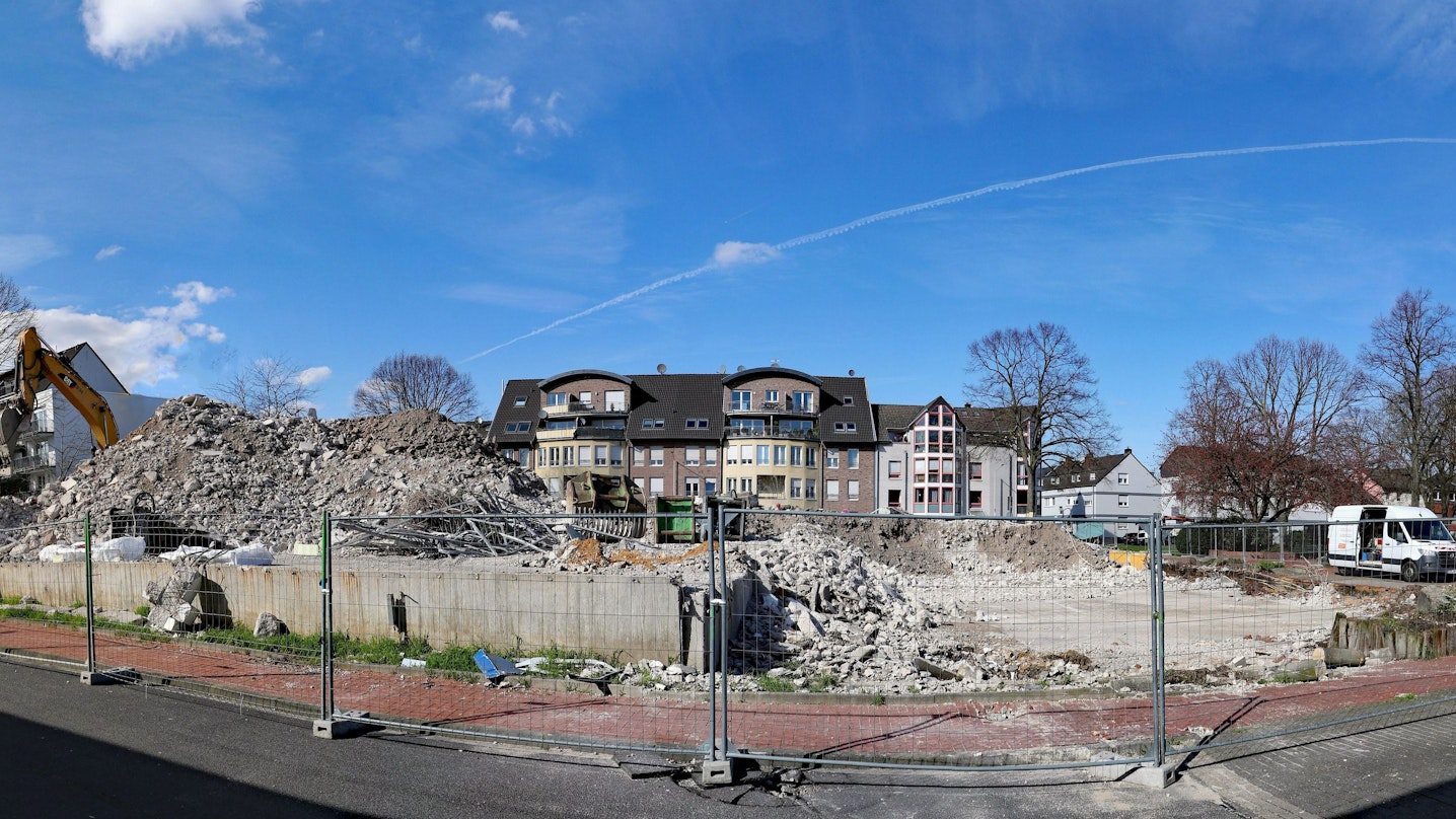 Hinter einem Zaun ist links ein riesiger Schuttberg zu suchen, dahinter stehen Wohnhäuser.