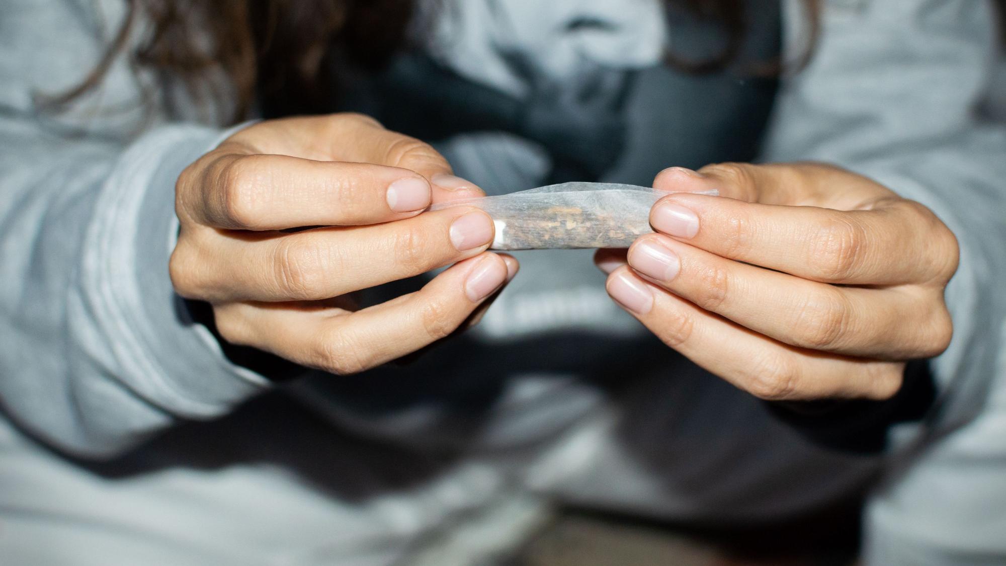 Eine junge Frau rollt nachts einen Marihuana-Joint in einer Straße. Zu sehen sind ihre Hände, in denen sie eine Cannabis-Zigarette hält.
