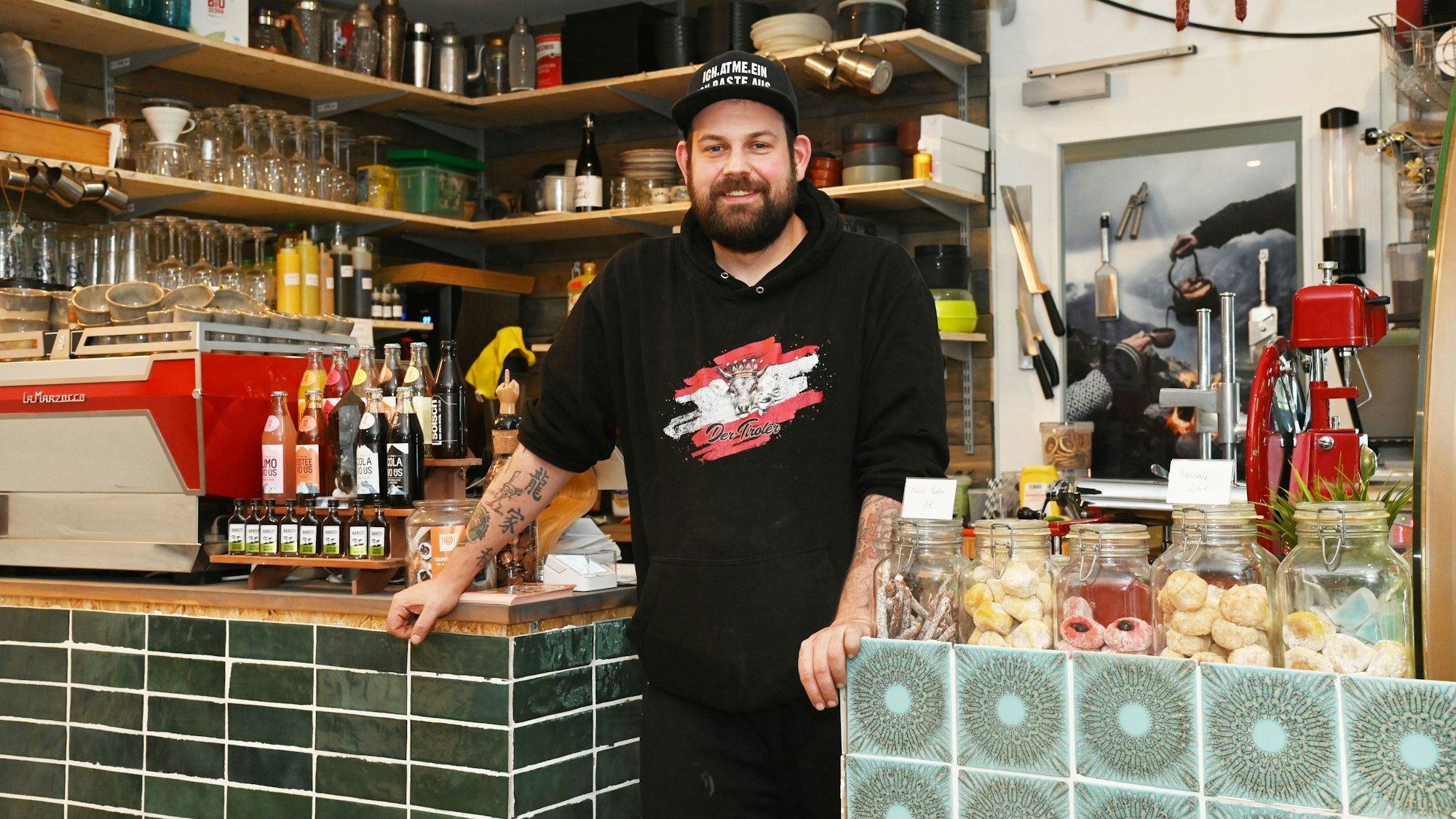 Ein Mann lehnt zwischen zwei Theken, er trägt einen schwarzen Pullover mit dem Logo seines Cafés und eine Kappe.