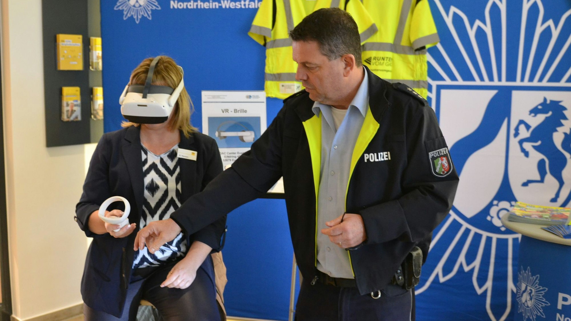 Eine Frau hat eine VR-Brille an, ein Mann in Polizeiuniform hilft ihr.