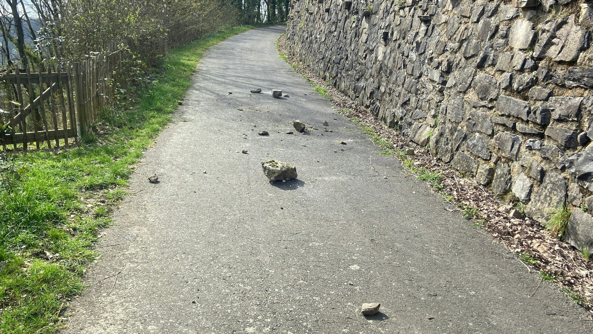 Unterschiedlich große Steine liegen auf einem asphaltierten Weg neben einer Naturscheinmauer.