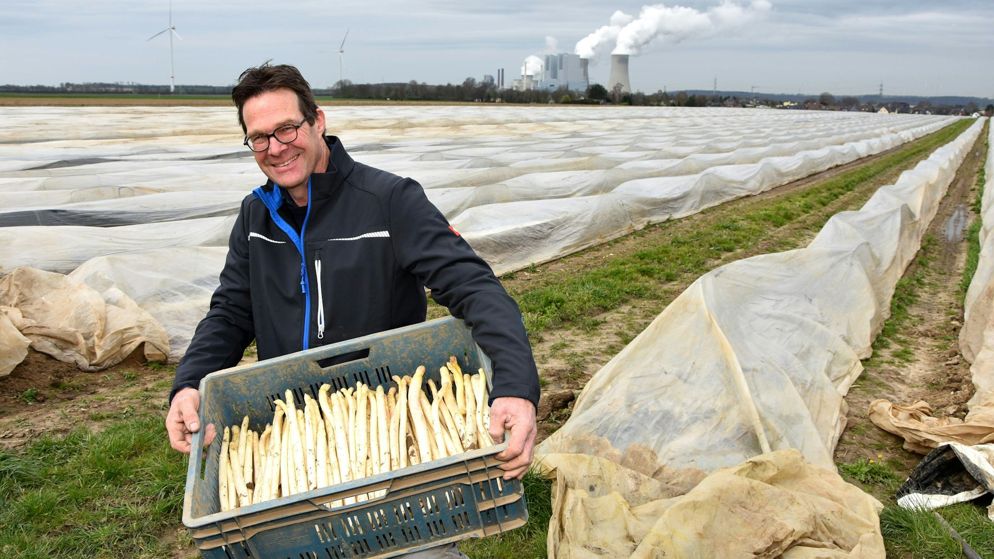 Ein Mann trägt eine Kiste Spargel von einem Feld, im Hintergrund steht ein Kraftwerk.