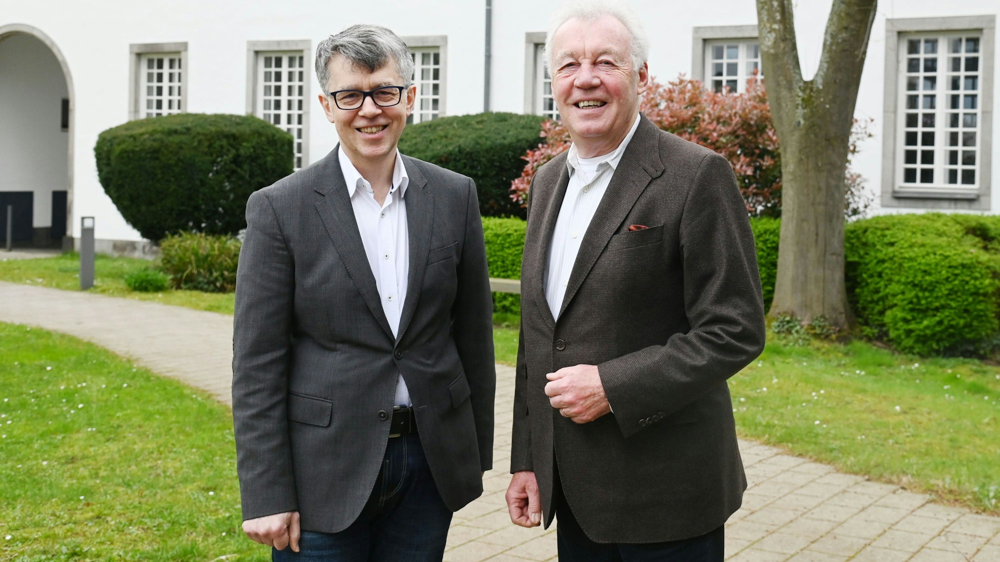 Zwei Männer in grauen Sakkos und weißem Hemd stehen vor einem Gebäude und lächeln in die Kamera.