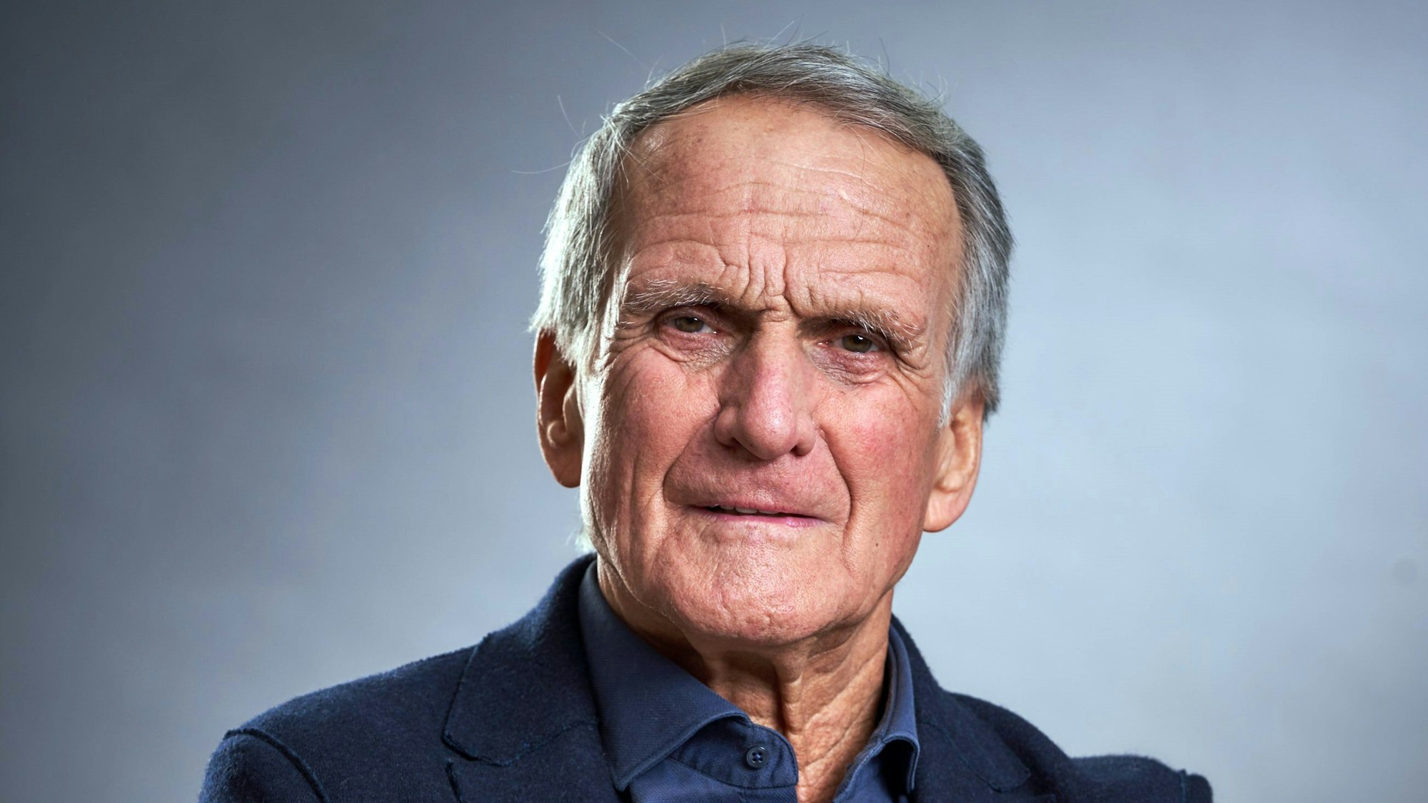 Wolfgang Overath schaut mit verschränkten Armen und gewohnt verkniffenem Blick in die Kamera.