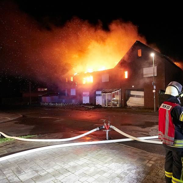 Mehrere Feuerwehrleute stehen vor einem brennenden Haus.