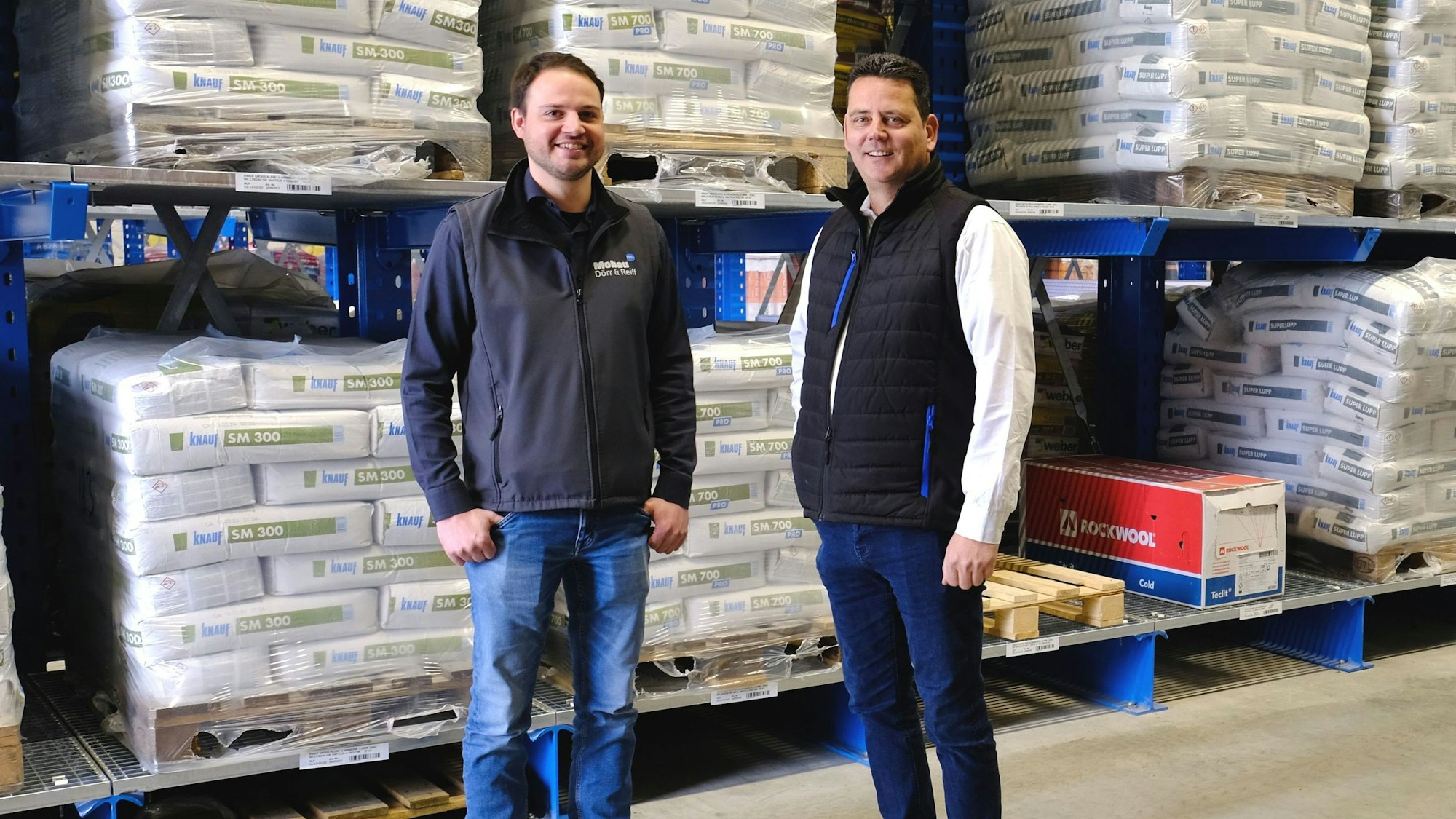 Dennis Runte (l.) und Dominik Kratz stehen in einem Lagerhaus vor einem Hochregal.