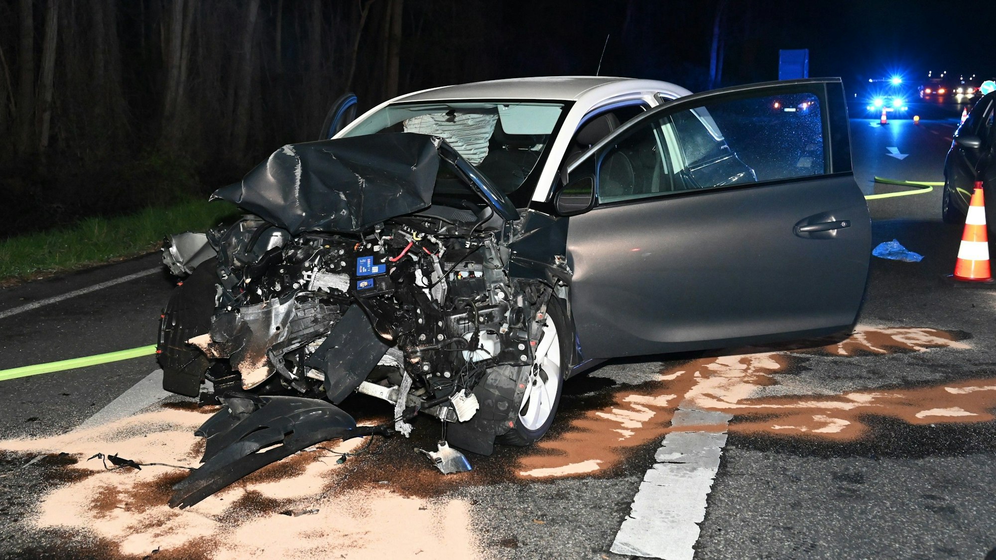Ein schwer beschädigter Pkw steht nach einem Frontalzusammenstoß auf einer ausgeleuchteten Straße in der Nacht.