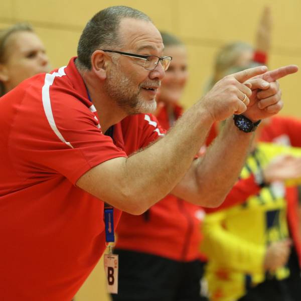 Andreas Kunzke deutet von der Trainerbank aus in Richtung Spielfeld. Im Hintergrund feiern mehrere Handballerinnen den Sieg ihrer Mannschaft.
