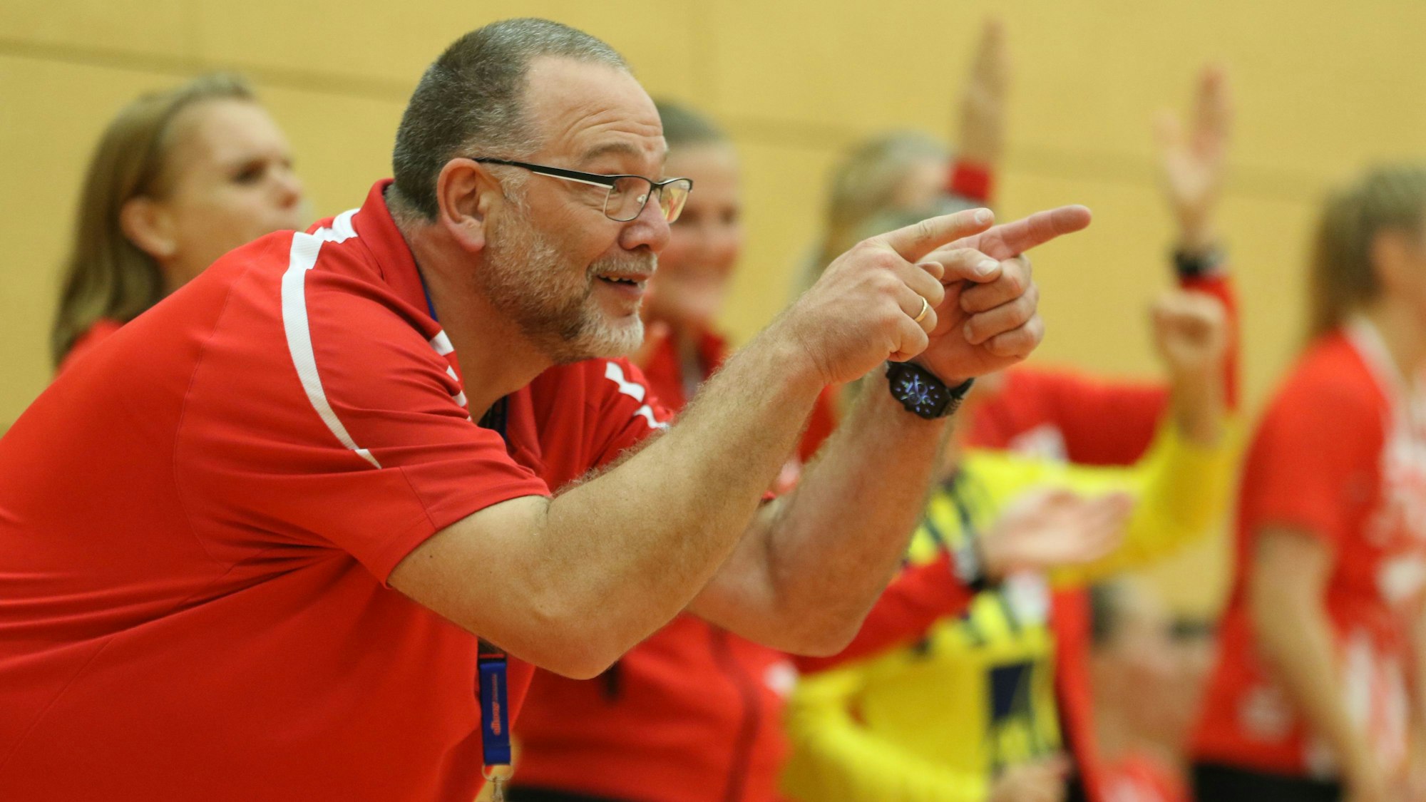 Andreas Kunzke deutet von der Trainerbank aus in Richtung Spielfeld. Im Hintergrund feiern mehrere Handballerinnen den Sieg ihrer Mannschaft.
